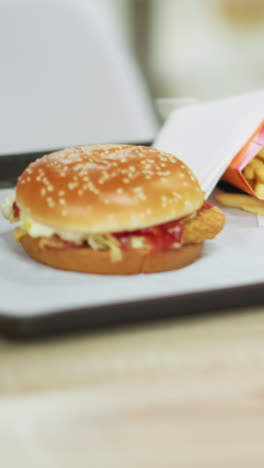 primer plano de las manos de una mujer en un vestido rosa dejando caer una bandeja negra con papas fritas, hamburguesa y café, ella ajusta la bandeja, alcanzando las papas frites, con una iluminación tenue y un fondo borroso