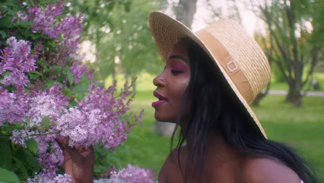 Mujer-Negra-Con-Vestido-De-Verano-Y-Sombrero-De-Paja-Huele-A-Flores-Lilas-Rosas-En-El-Parque,-Sonríe-Ante-El-Olor-Agradable---Cierra-A-Cámara-Lenta