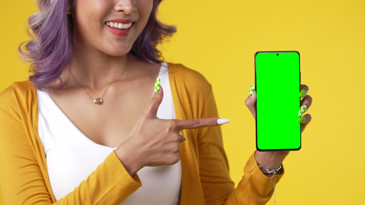 joven asiática feliz mostrando un teléfono inteligente con pantalla verde aislada sobre un fondo amarillo