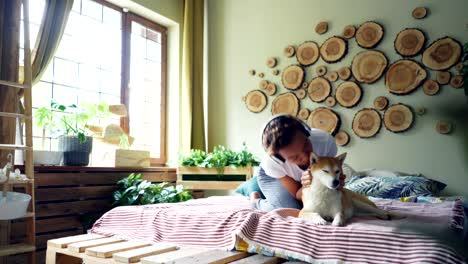 cute mixed race woman is listening to music with headphones, dancing sitting on bed and stroking her adorable shiba inu dog with love and care. people, technology and animals concept.