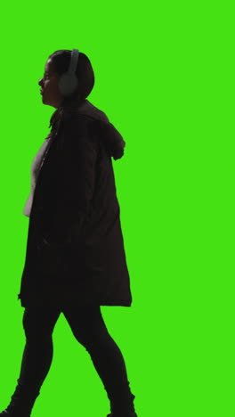 vertical video full length shot of woman wearing wireless headphones streaming music from mobile phone walking across frame against green screen 2