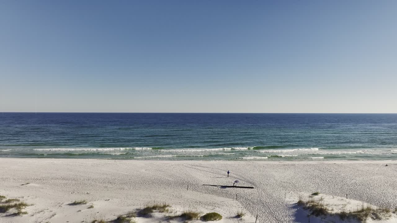 Push-in wide aerial drone shot of a man walking towards the sea or the ocean on an empty white sand beach, with clear blue sky and horizon, capturing the serene atmosphere and expansive coastline