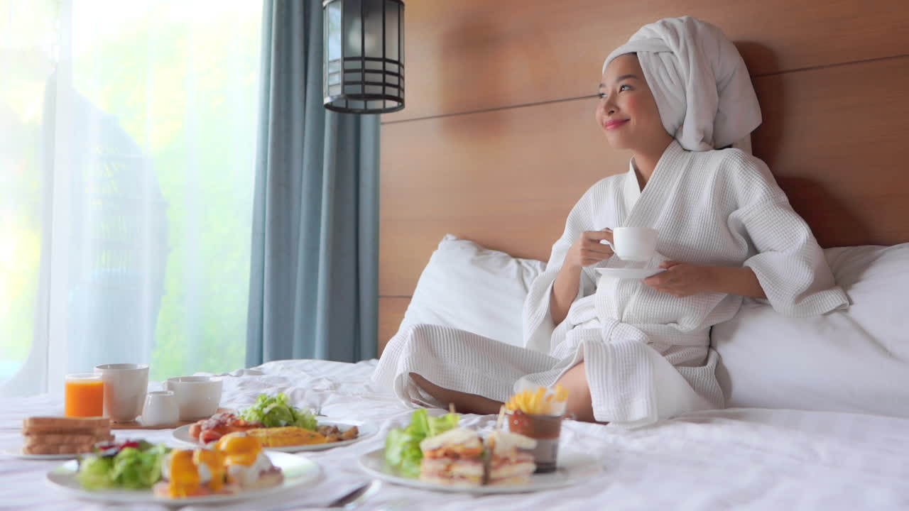 Static shot of a sexy Asian lady having breakfast and coffee in a hotel bed while wearing a robe and a towel on her head