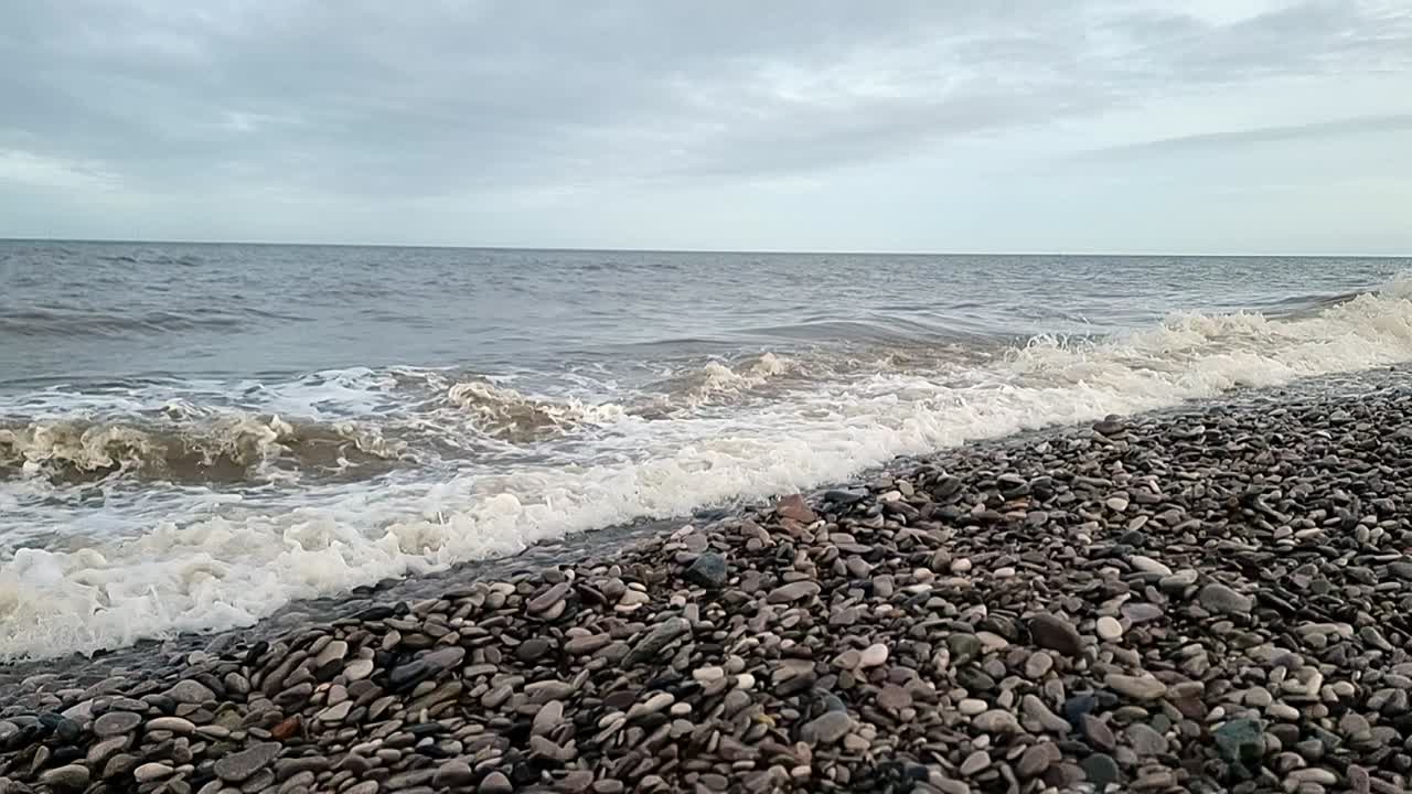 바람이 불어오는 가을에 웨일즈의 자갈 해변에 느린 속도로 부히는 모래 바다 파도