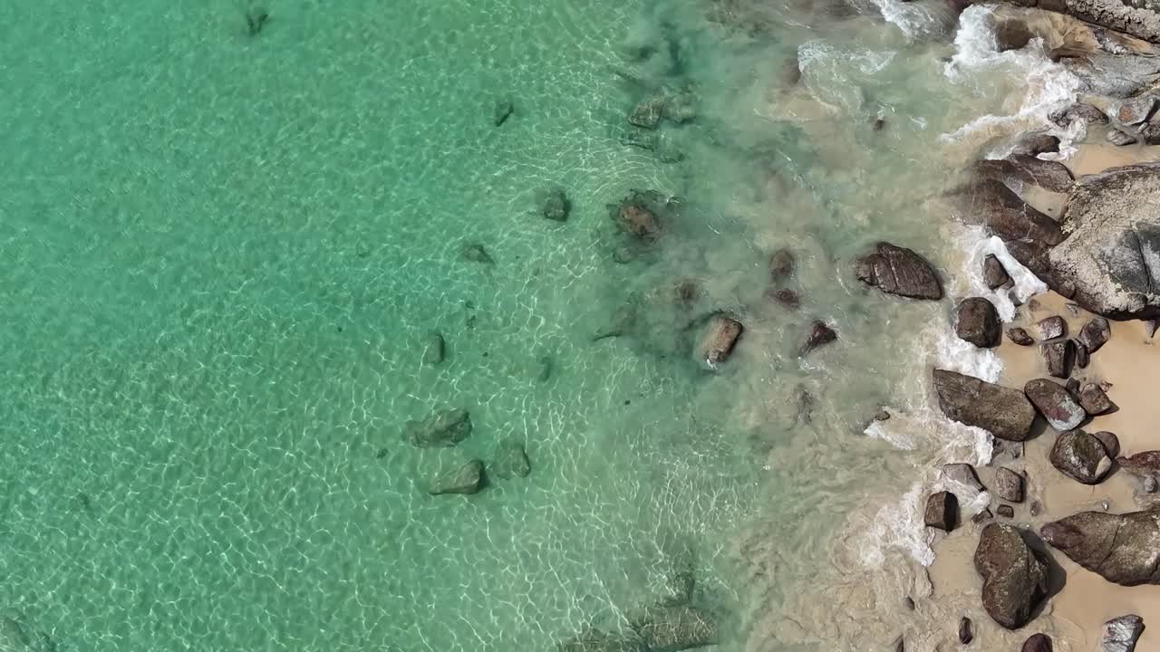Aerial view of clear turquoise waters meeting a rocky shoreline with gentle waves and sandy beach.
