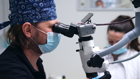 doctor in mask and cap looking attentively at the advanced microscope. healthcare specialist saying something to the nurse sitting beside.