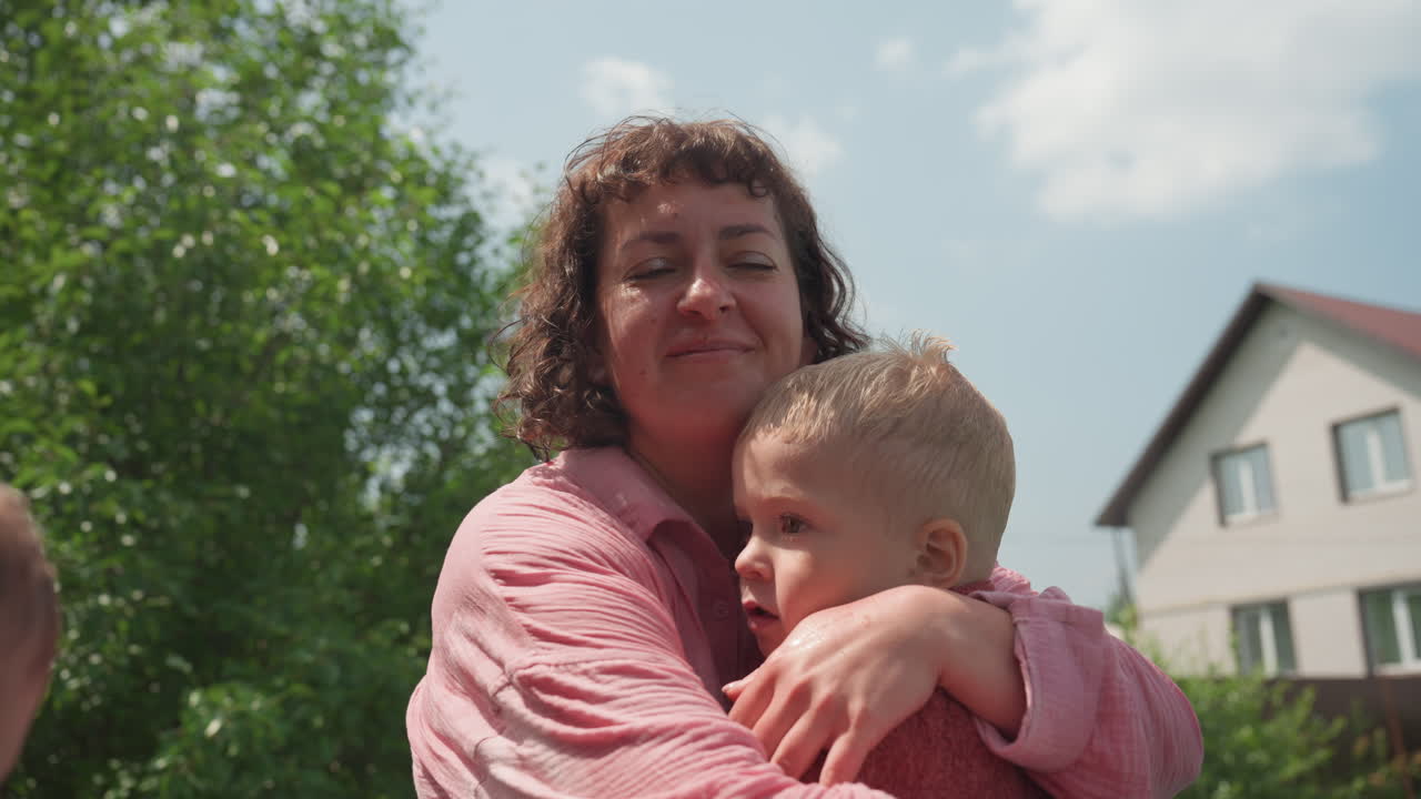 Mutter umarmt Kind, kaukasische Mutter hält ihr Kind sanft in einer hellen Gartenumgebung. Frau stellt zärtliche, schützende Umarmung mit ihrem Nachwuchs in üppiger Außenumgebung in der Nähe ihres Wohnsitzes dar