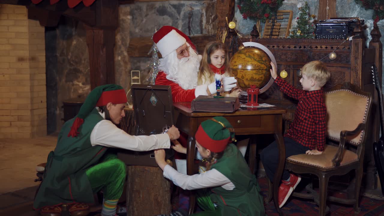 Santa, little kids and two elves in Santa's residence. Elves in green costumes playing with old recorder. A boy and a girl looking at globe together with Santa.