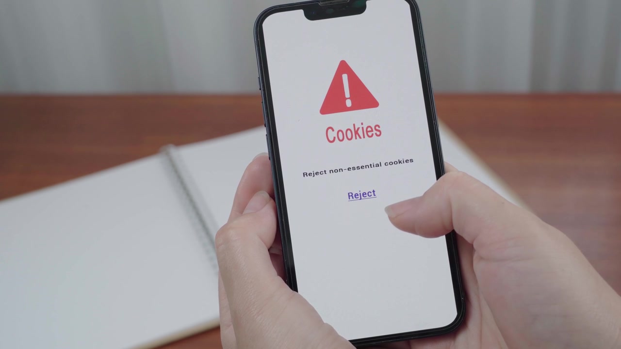 User holds a smartphone displaying a cookie consent warning screen and moves a thumb toward the reject non essential cookies option.