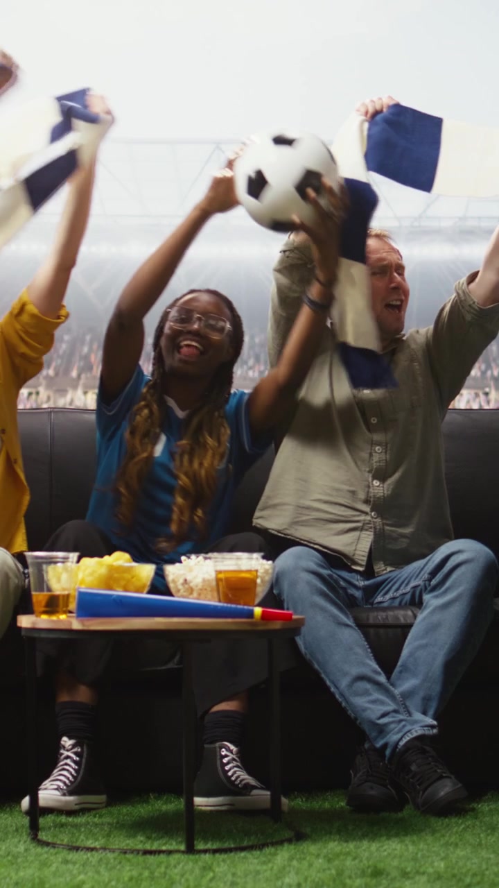 Video vertical: Aficionados al fútbol en el sofá de casa con bufandas de su equipo viendo un partido inmersivo.