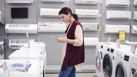 Young-brunette-picks-up-a-pricetag-from-the-top-of-washing-mashine-in-appliance-store.-Looking-for-perfect-household-equipment.