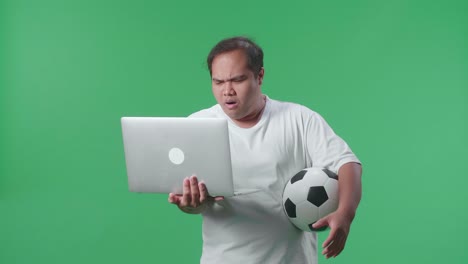 un hombre asiático con una pelota se arrepiente de animar al fútbol en una computadora portátil en el estudio de pantalla verde