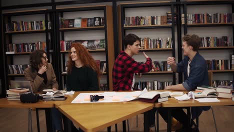 slow motion of students making fun after hours of studying in uni library. happy and free students - two men greeting each other, gesturing. two girls discussing something sitting at the table in library