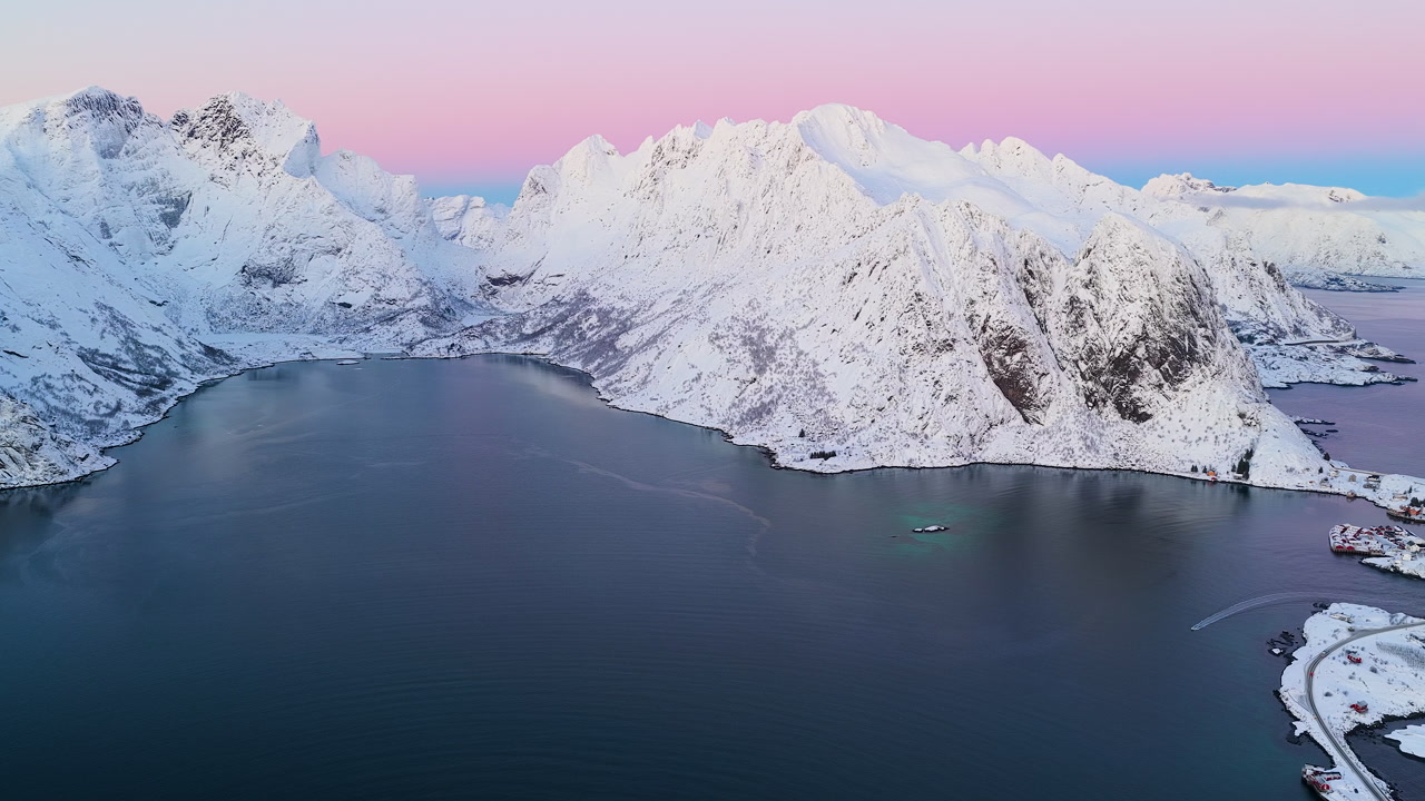 Stunning aerial panorama of snowcapped peaks surrounding a calm fjord in Lofoten, Norway during a vibrant pink winter sunset.