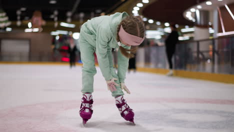 a close view of a child in a mint green outfit and pink skates bending down to clean herself on an ice rink, other skaters can be seen in the blurred background