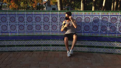 a man wearing a cloth mask walks up and sits down on a tiled park bench then removes his mask to enjoy a coffee break in the sun