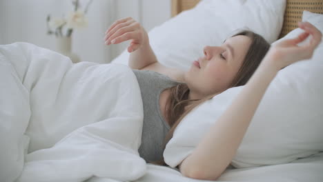 beautiful brunette is waking up in the morning stretches in the bed sun shines on her from the big window. happy young girl greets new day with warm sunlight flare.