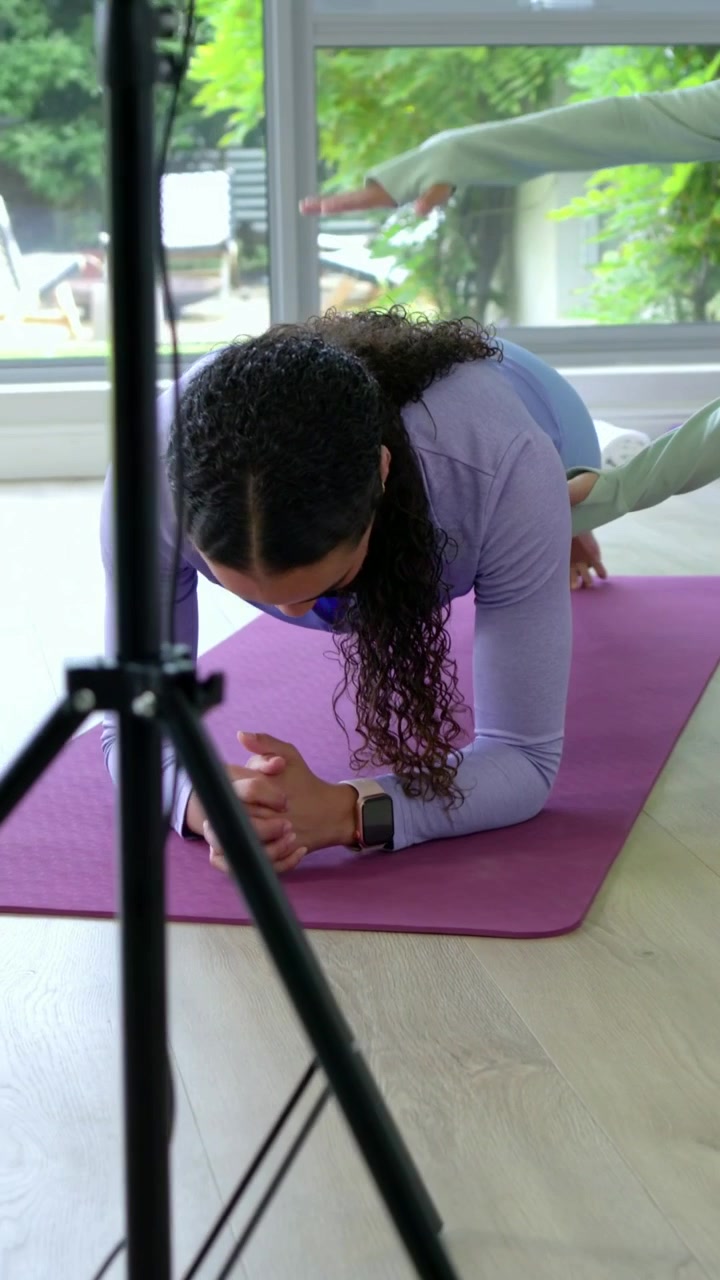 Video vertical: Entrenador dando instrucciones a una mujer con top lila, activando los abdominales y haciendo una plancha en una colchoneta en casa para mejorar la postura.