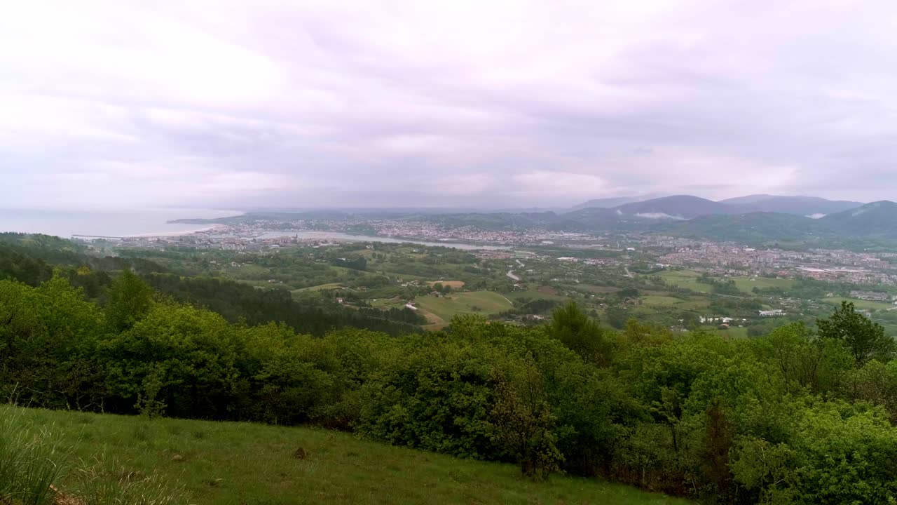 Panoramic view of Irun, Spain, with the Bay of Biscay, green hills, and distant misty mountains under an overcast sky