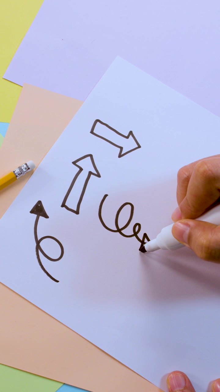 Person draws various black arrows and curly symbols on white paper, overhead view, bright lighting