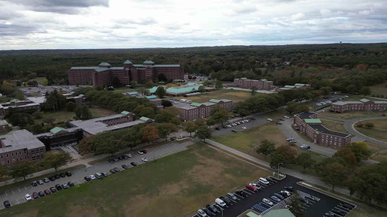 Aerial drone view of a large medical campus in Brentwood, Suffolk County, Long Island, NY, showcasing multi-wing architecture, structured parking, open grounds, and surrounding landscape in clear day