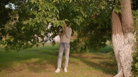 woman in casual attire and white sneakers stands under tree, focused on phone screen with hand touching hood collar, sunlight filtering through leaves casting soft reflection on her jeans