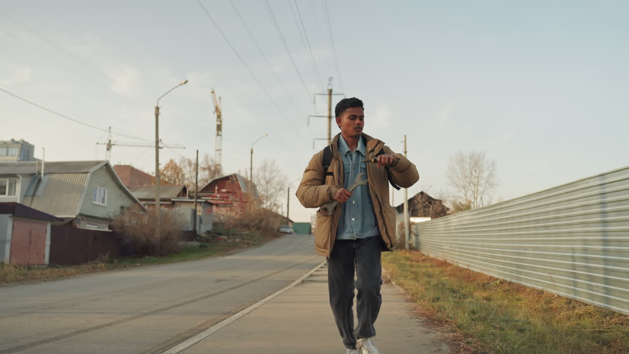 Tecnico di riparazione asiatico che cammina verso la telecamera su una strada suburbana, a passo deciso con zaino e strati di denim, recinzione ondulata e pali dell'elettricità che incorniciano la scena, bagliore del tardo pomeriggio, approccio determinato.