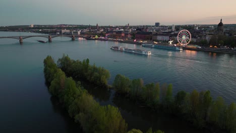 Vuelo-De-Drones-Al-Atardecer-De-Mainz-Sobre-El-Río-Rin-Cruzando-Un-Barco-Con-Un-Festival-De-Vino-En-La-Orilla-Del-Río-Y-El-Cielo-De-La-Tarde-De-Verano-En-La-Parte-De-Atrás