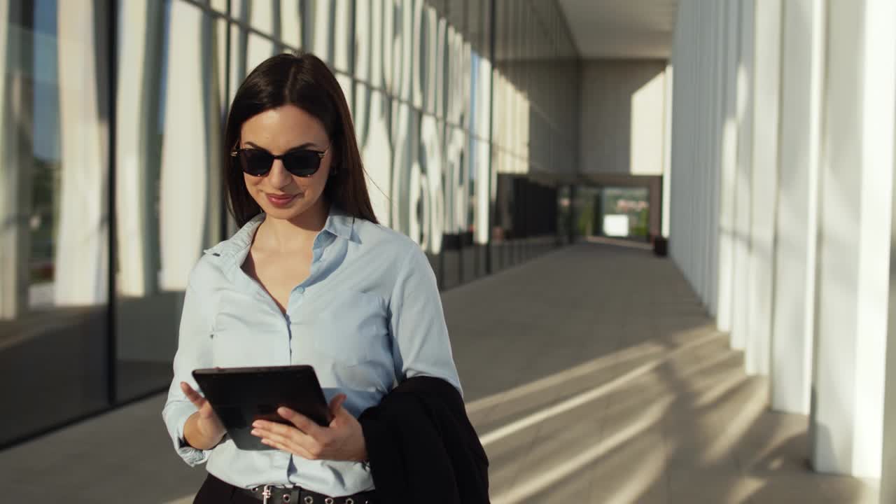 mujer de negocios caminando al aire libre frente al edificio de oficinas y usando una tableta digital