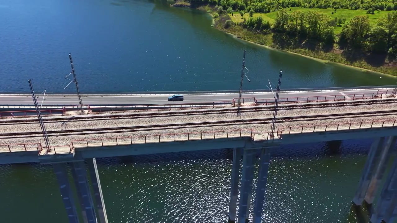 Вид на автомобильный и железнодорожный мост, пересекающий реку, с автомобилем, едущим вдоль воды, в ясный солнечный день, демонстрирующий зелень и инфраструктуру.