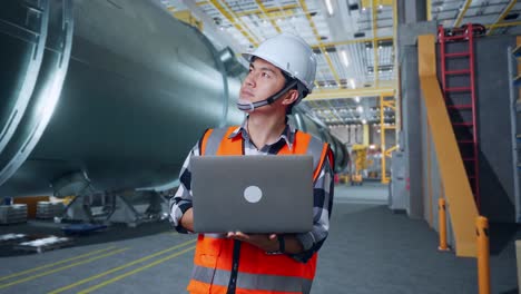 ingeniero masculino asiático con casco de seguridad mirando una computadora portátil y mirando a su alrededor mientras está de pie en una fábrica de fabricación de tubos