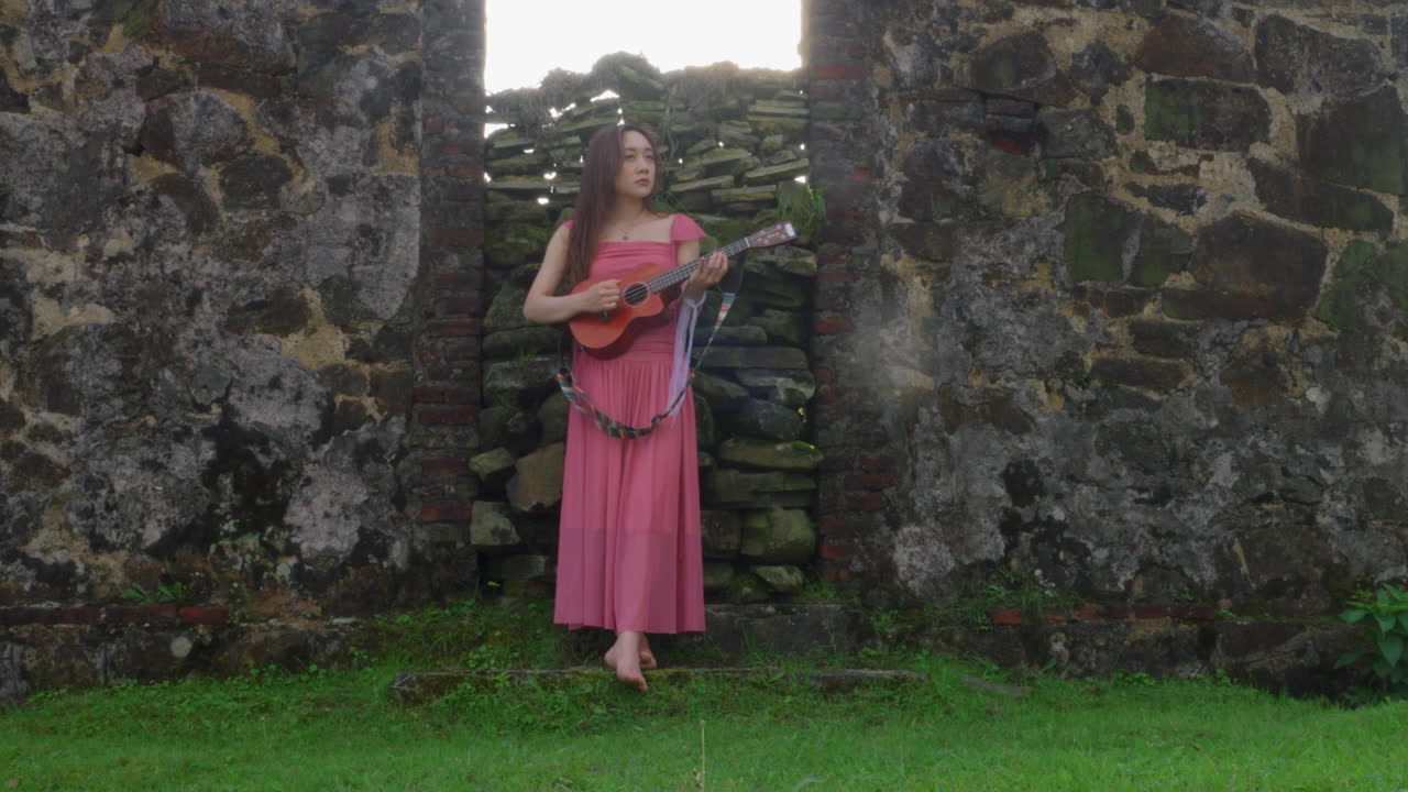 young asiatic artist playing guitar ukulele outdoor in nature wearing dress red