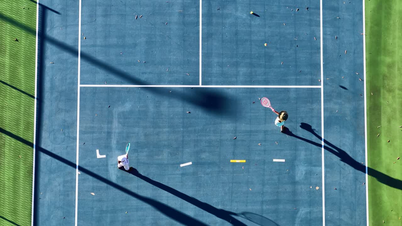 Two players engage in a tennis match on a sunlit court, captured from a drone perspective