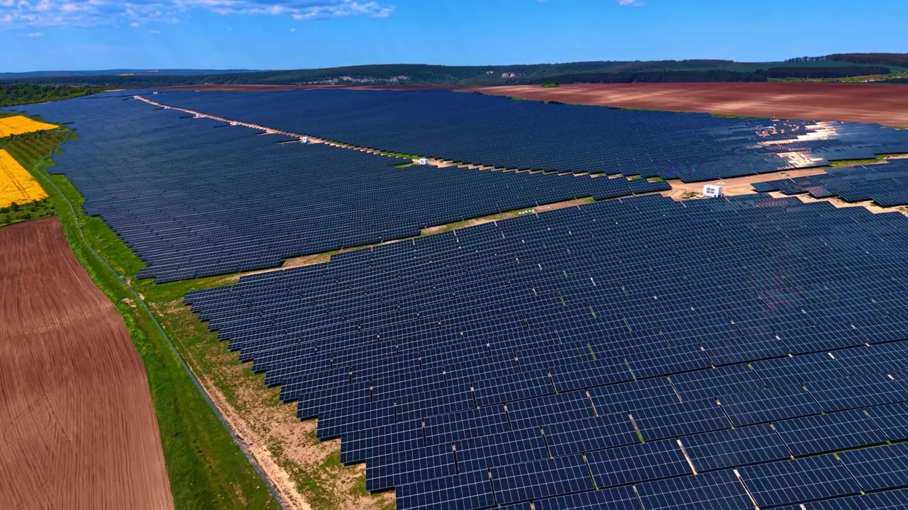Massivt solcelleanlæg omgivet af landbrugsjord og rapsmarker. Fladt terræn med mørke panelrækker, pløjet brun jord og gule afgrødearealer under sommerhimmel.