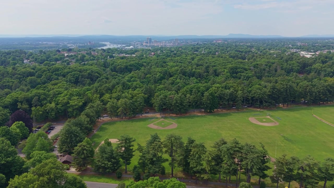 Vol de drone incliné vers le haut au-dessus de vastes terrains de baseball entourés de bois dense dans une zone de loisirs communautaire avec l'horizon lointain de Springfield, Massachusetts, États-Unis