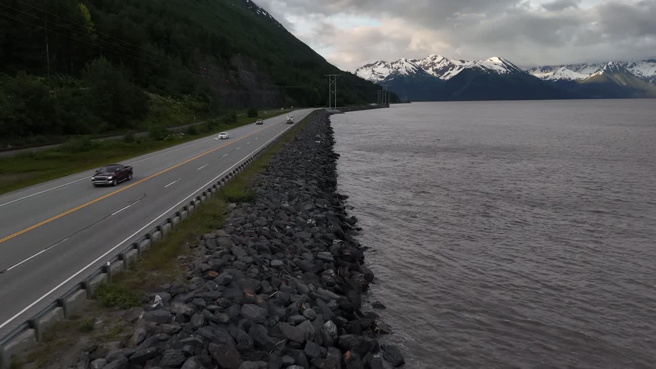 Aerial View of cars driving down road, early morning in late June along Seward Highway in Alaska, Drone flying over Rocky shore along Turnagain Arm, Snow covered mountains in background,