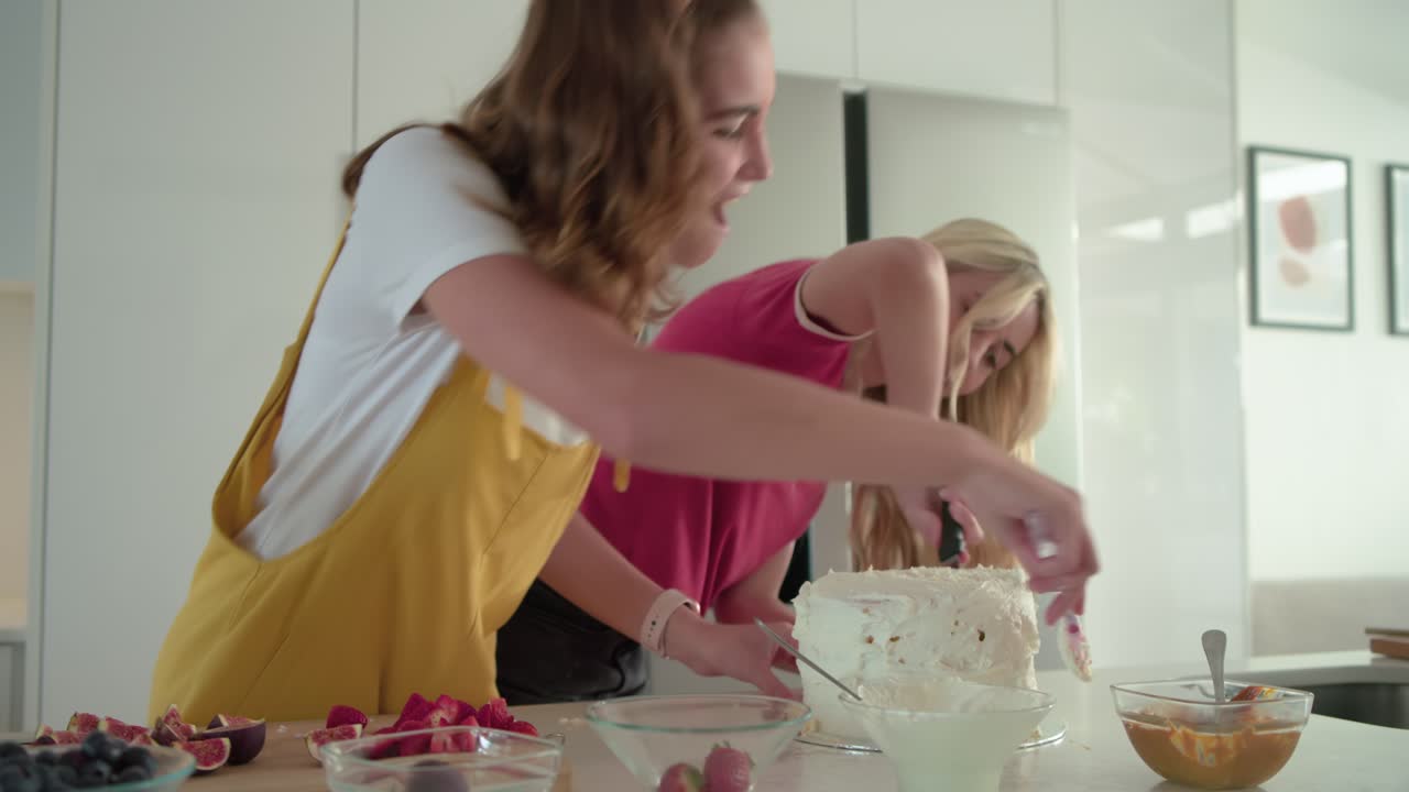 Two female friends are scraping frosting, sparking spatula tugging, then dancing on kitchen island