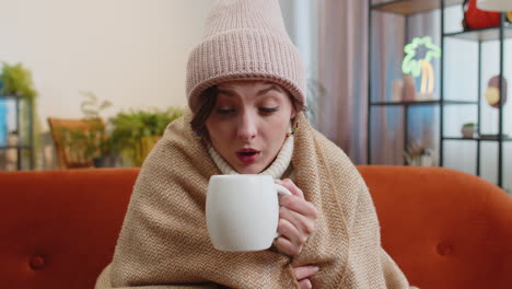 sick woman wear hat wrapped in plaid sit alone shivering from cold on sofa drinking hot tea