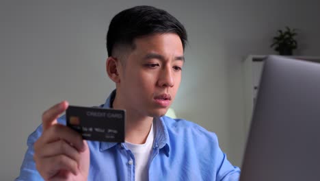 serious young asian man, frowning and worried while using and looking at credit card, doing online shopping, making online payment transaction using laptop at home.