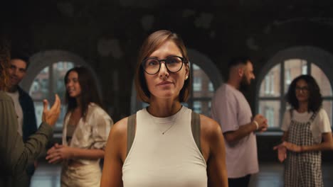 a girl with a bob in round glasses in a white t-shirt posing and looking at the camera against of group therapy in a brick hall. happy group therapy participant