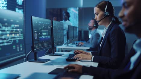 feliz y confiado especialista en soporte técnico al cliente que tiene una llamada de auricular mientras trabaja en una computadora en una habitación de monitoreo y control oscura llena de colegas y pantallas de visualización.
