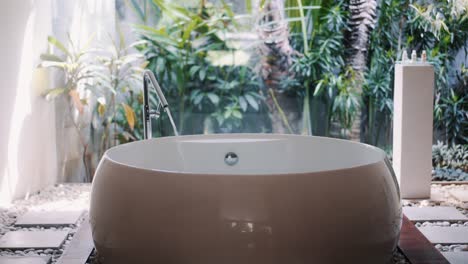 water pours into round bathtub, luxury bathroom, tropical greenery on background