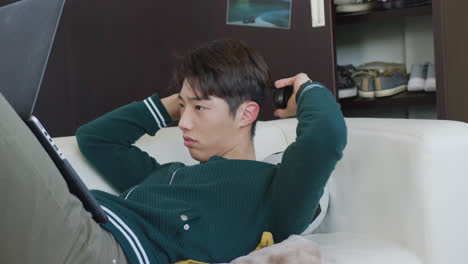 헤드폰을 착용 한 아시아 소년 이 집 의 소파 에 누워 있는 노트북 을 사용 하고 있다