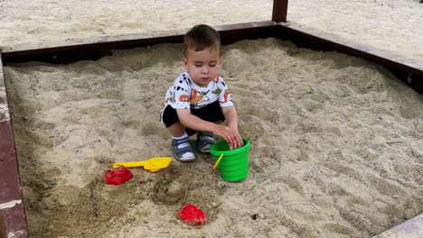 cute caucasian toddler boy sits squatting in the sand box. kid takes a handful of sand, pulls it to his mouth and then puts into bucket.