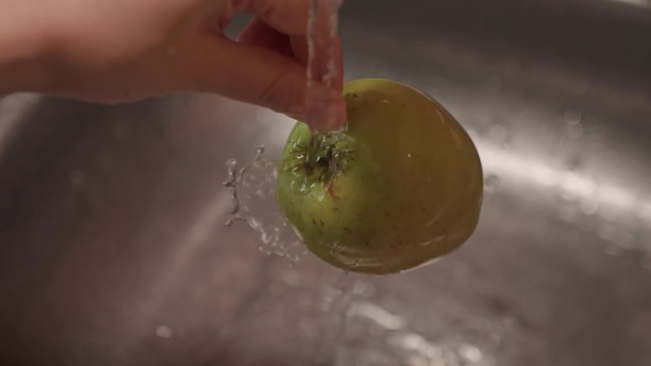 Mano sostiene manzana verde bajo el agua corriente, lavándola en el fregadero. Fluye un chorro de agua. Grifo de cocina de acero inoxidable con escurridor metálico y fregadero para lavar la manzana.