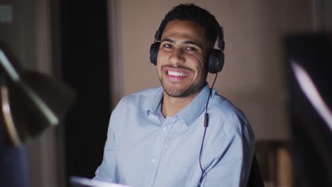 Video-portrait-of-smiling-biracial-businessman-wearing-headphones,-working-at-night-in-office