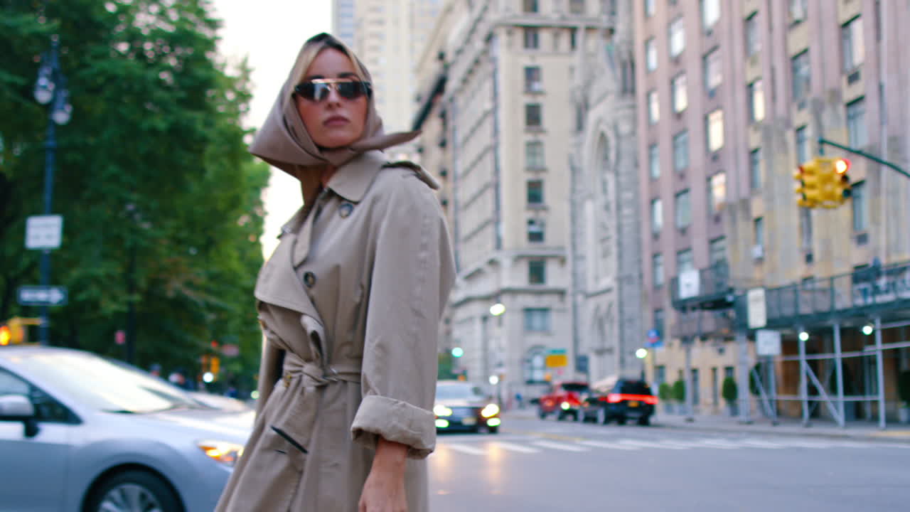 Donna in trench e foulard che attraversa una strada di New York. Donna alla moda che cammina su un passaggio pedonale cittadino indossando un trench beige e un foulard di seta al crepuscolo.