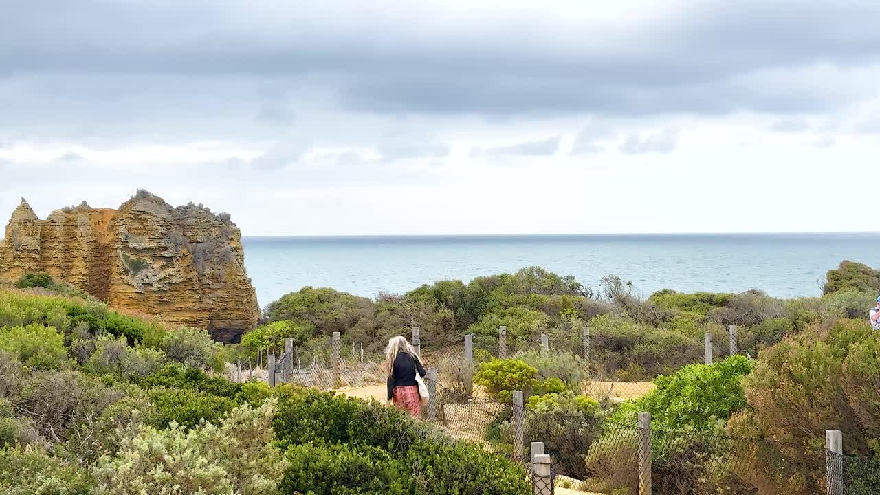 A woman walks through lush greenery on a coastal path with rocky formations and ocean views under cloudy skies