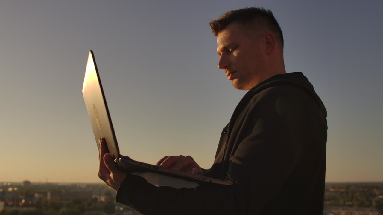 Programmer a hacker is on the roof with a laptop at sunset says error code on the keyboard and looking at the city view.