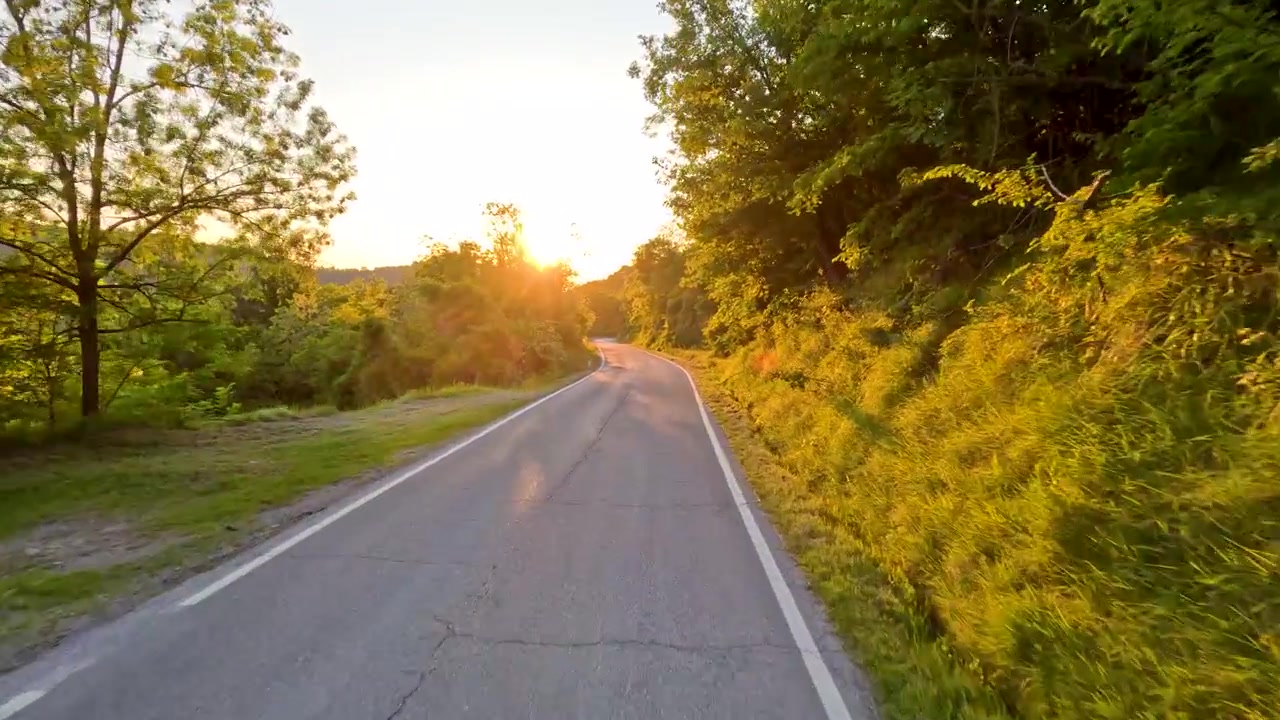 Prospettiva in prima persona mentre si guida lungo una strada asfaltata curva durante un vivace tramonto estivo.